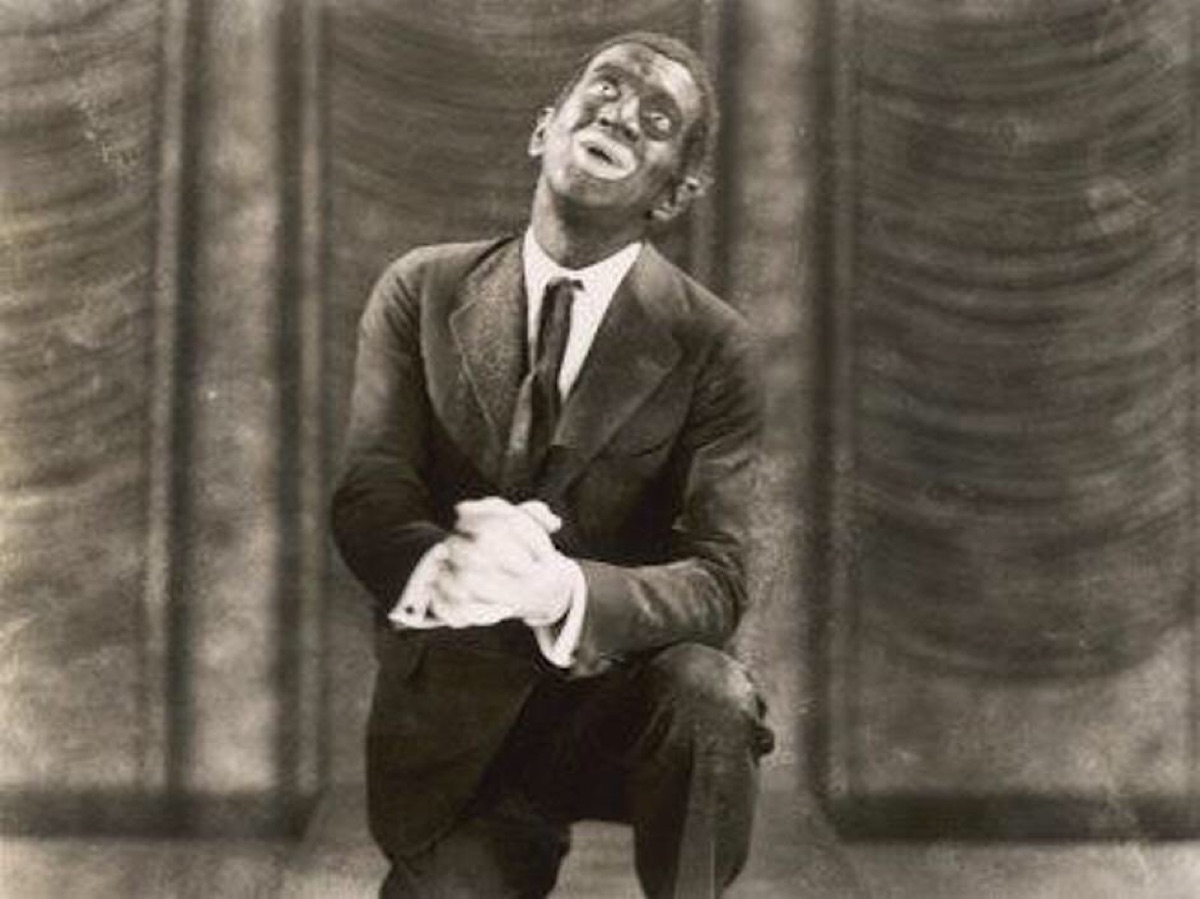 Al Jolson in blackface, kneeling on a stage with his gloved hands clasped