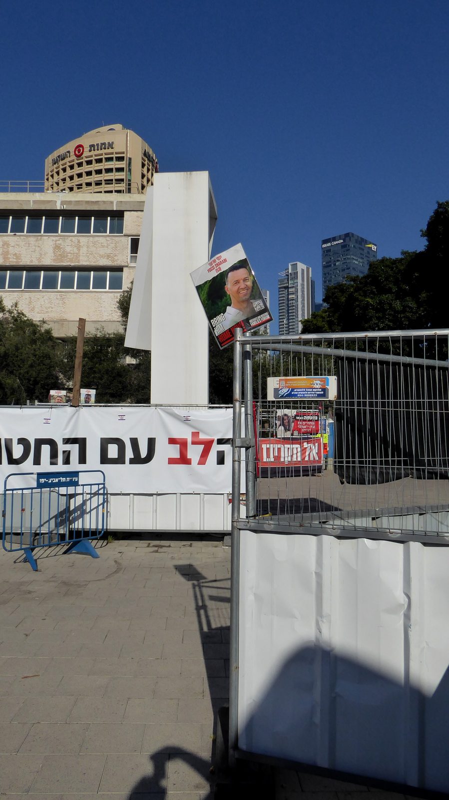 Open square entrance, banner and posters on fencing, skyscraper behind, Tel Aviv