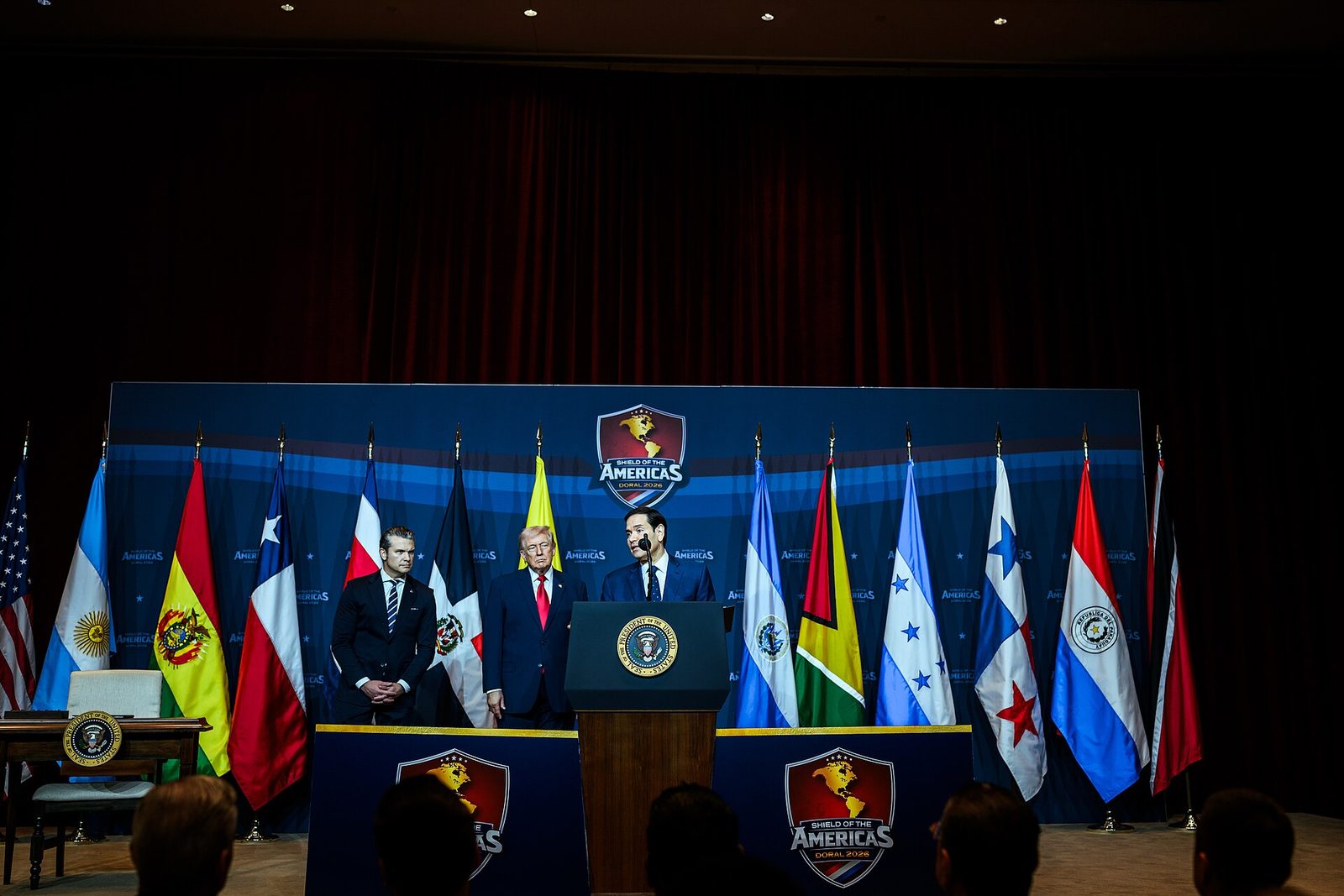 Wide stage shot with podium, row of national flags, Shield of the, Trump National Doral