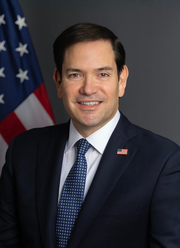 Smiling man in suit, US flag, grey studio backdrop.