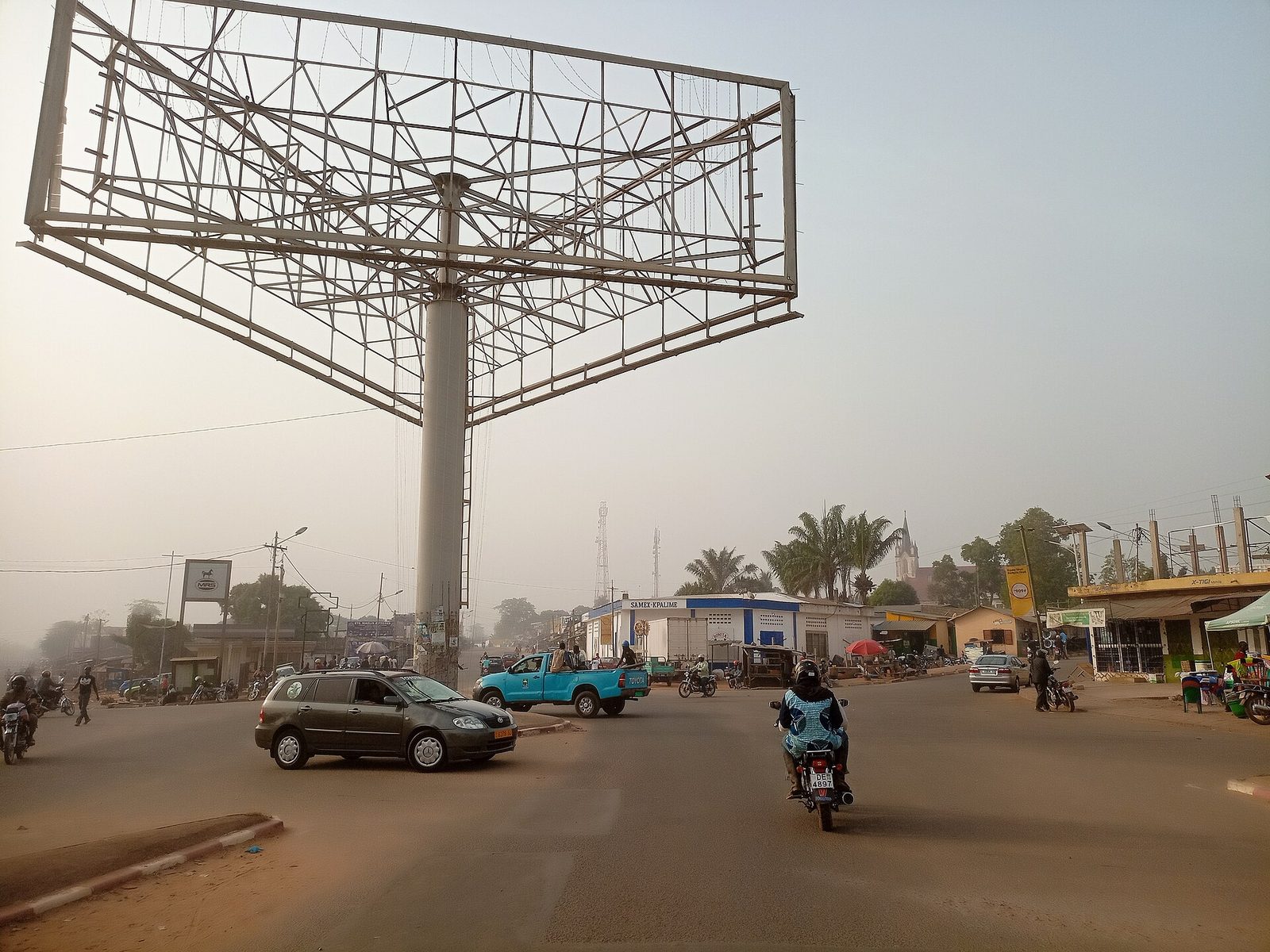 large town crossroads with metal billboard, vehicles and motorbikes, Kpalimé