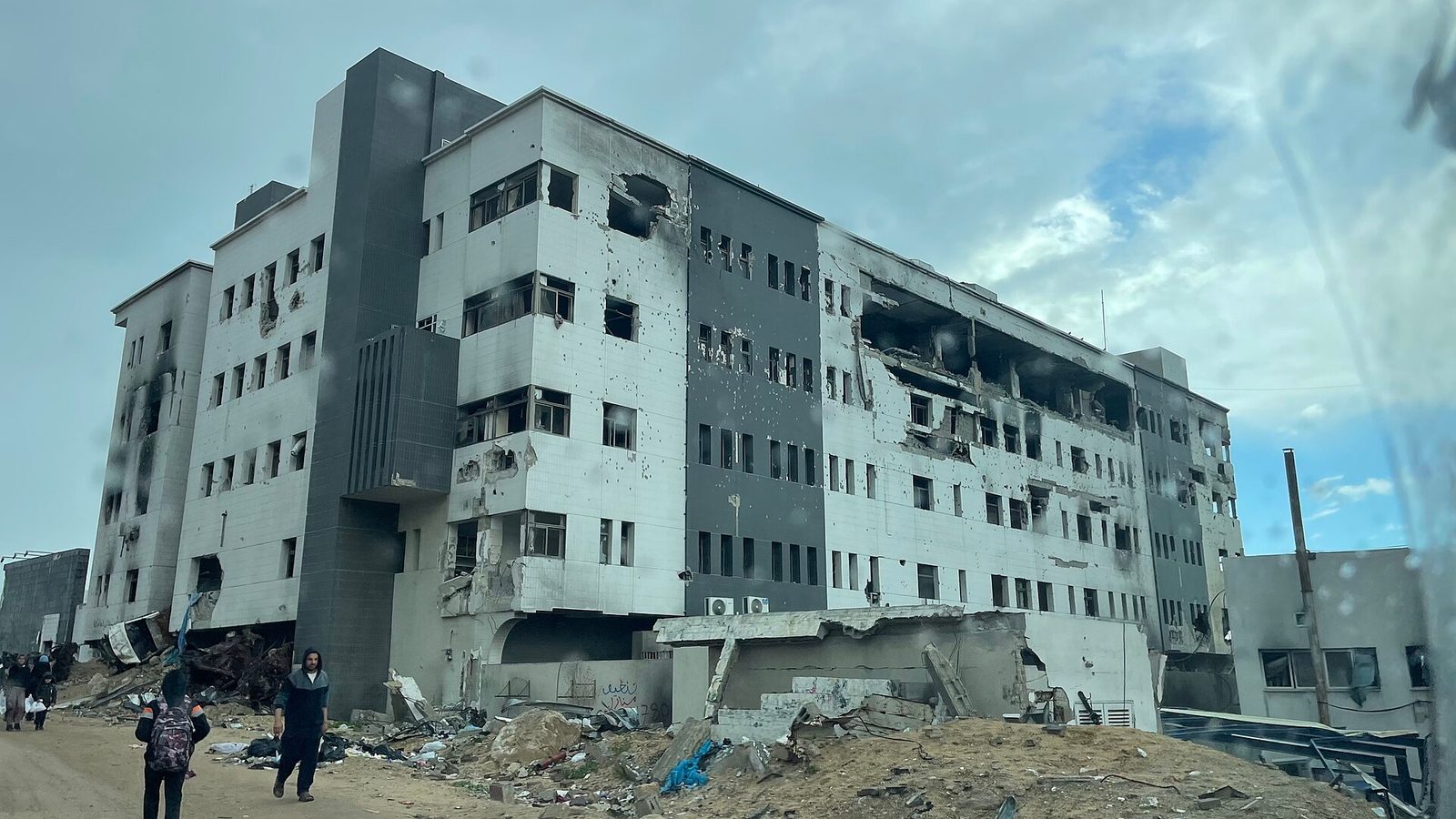 Large damaged hospital building, pockmarked walls, people walking past rubble