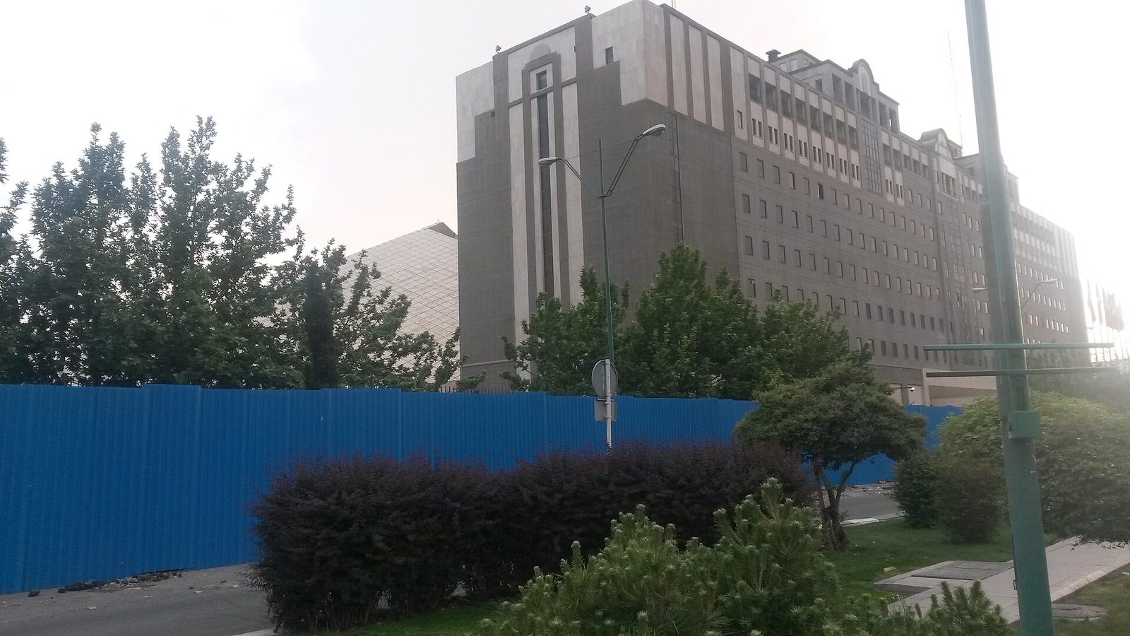 large concrete parliamentary building behind blue hoarding, trees, Tehran