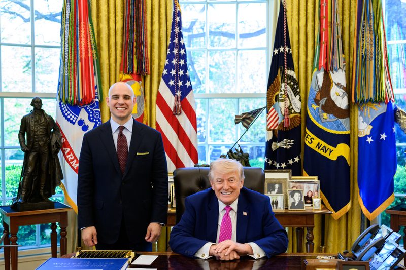Two men in an office with desk, presidential flags and window