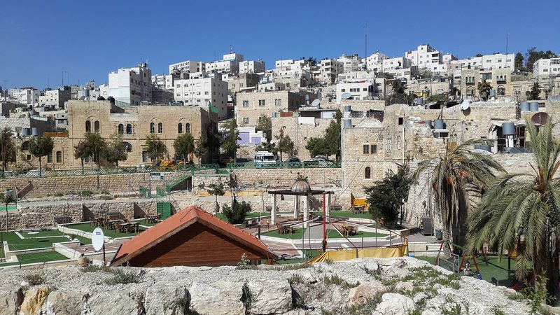 Stone buildings on a terraced hillside, courtyard and palm tree visible, Hebron