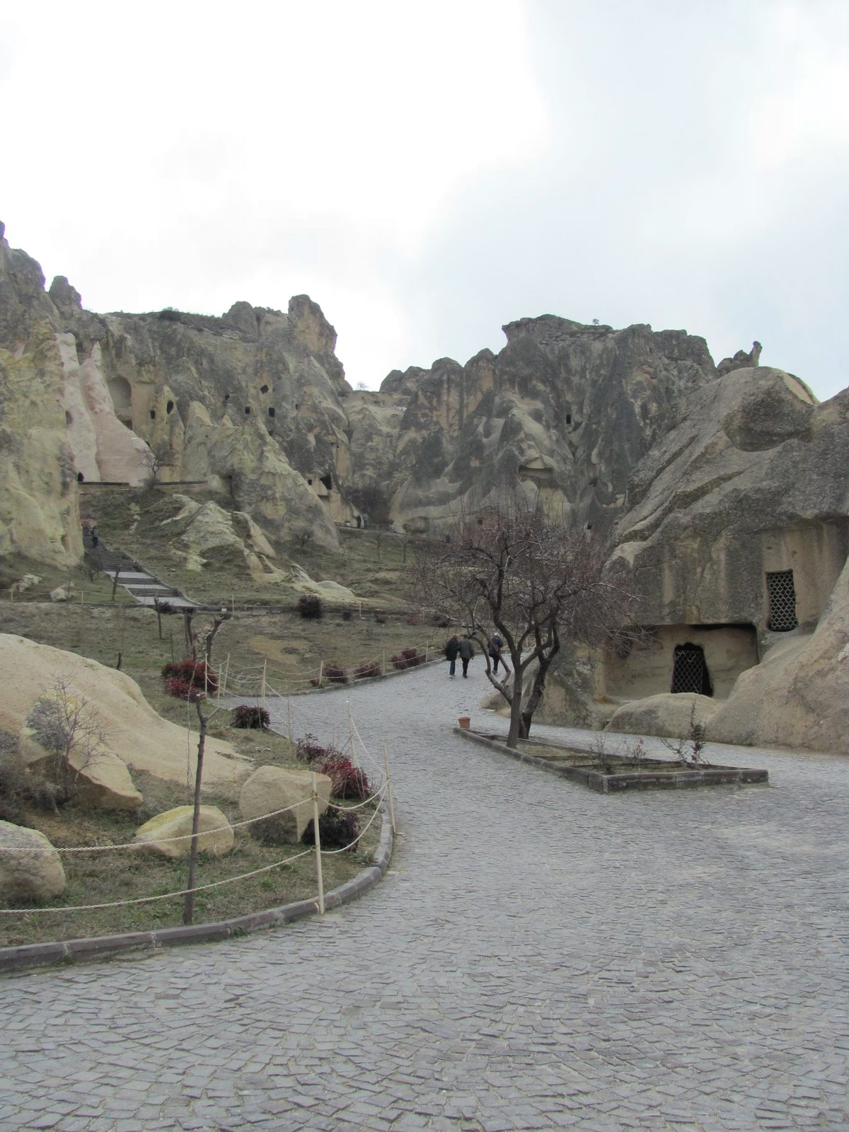Paved path through rock formations, carved openings, a few people walking, Goreme Open Air Museum