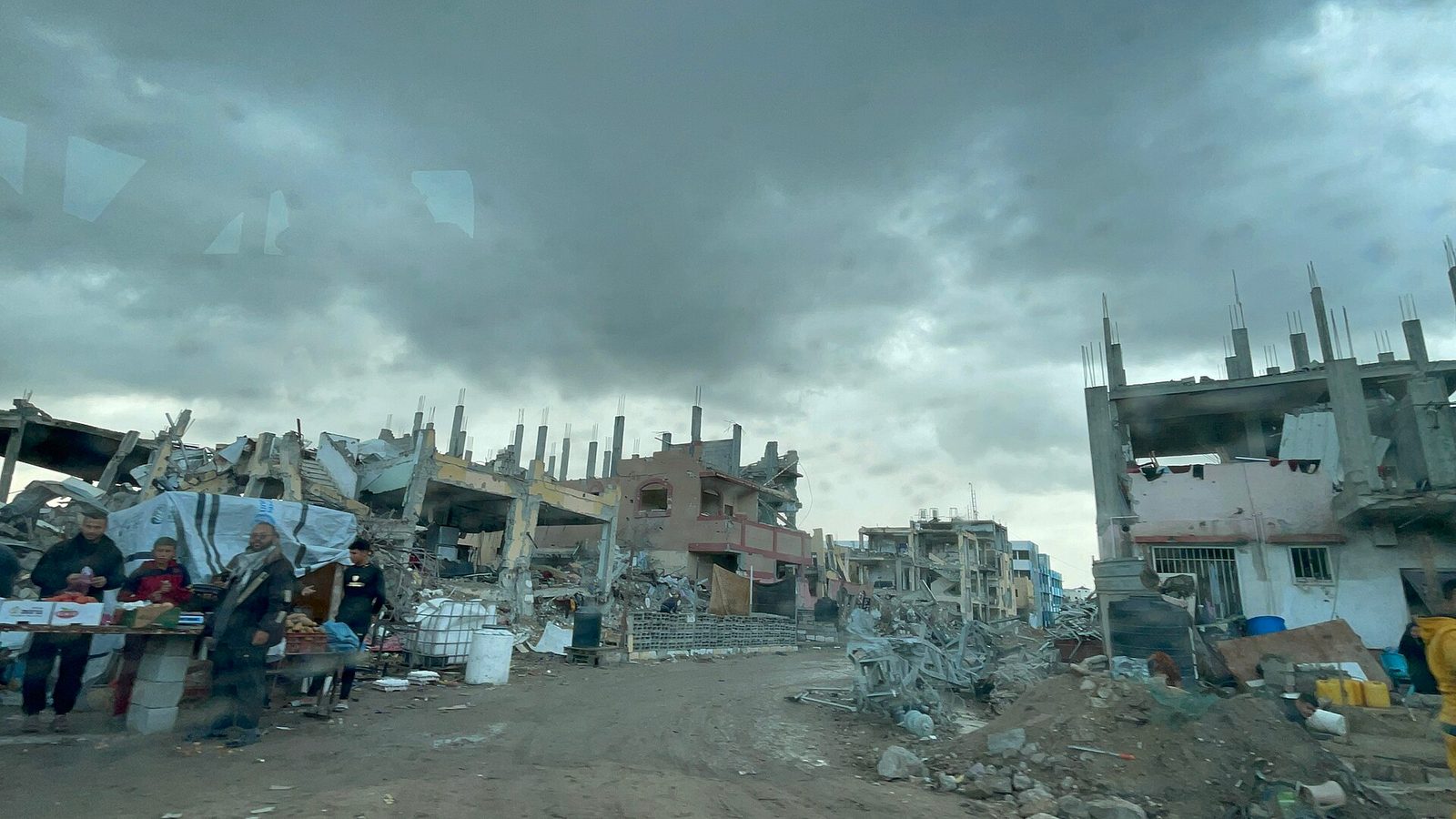muddy street lined with partially destroyed buildings, people and market stalls, Tel al-Sultan