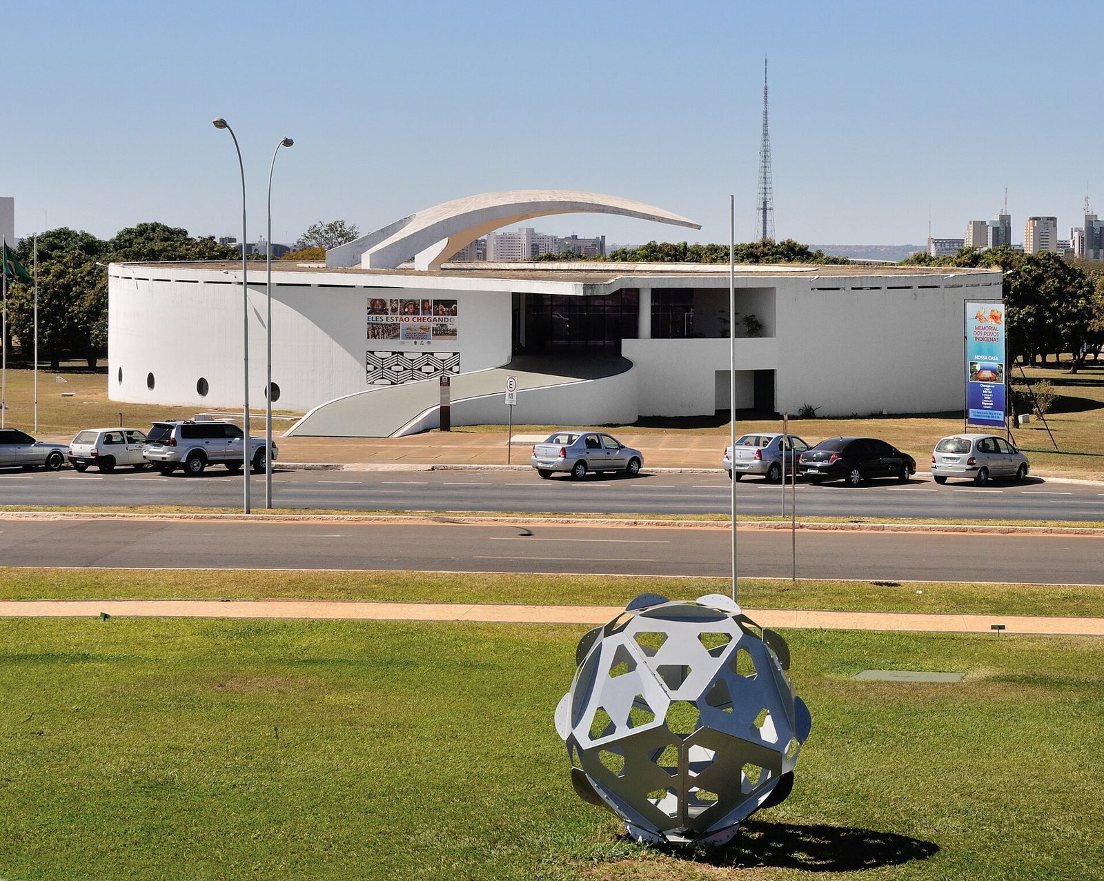 White curved memorial building on grass, parked cars on road, Brasília