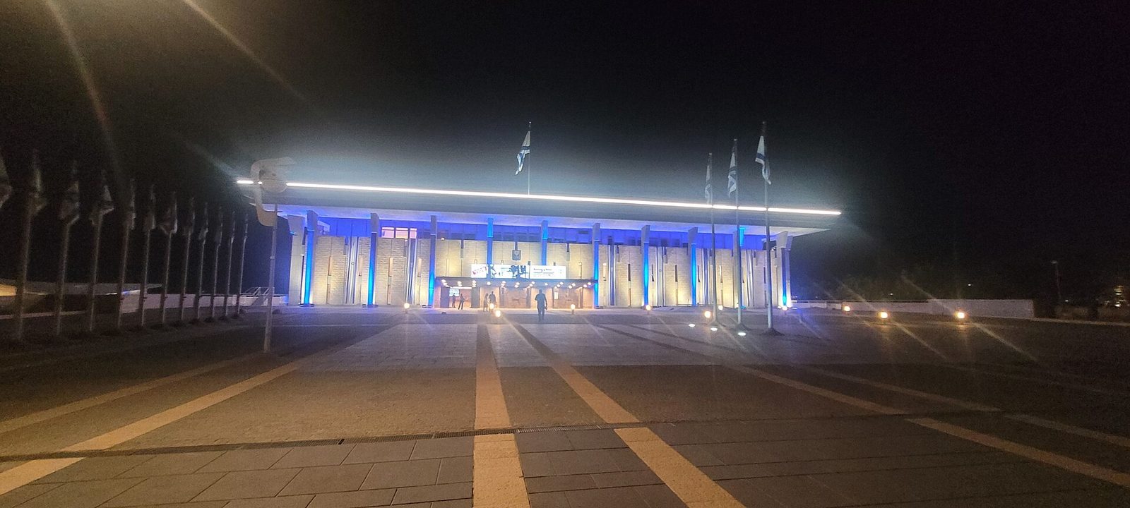 Knesset building lit at night, flags on poles, empty forecourt