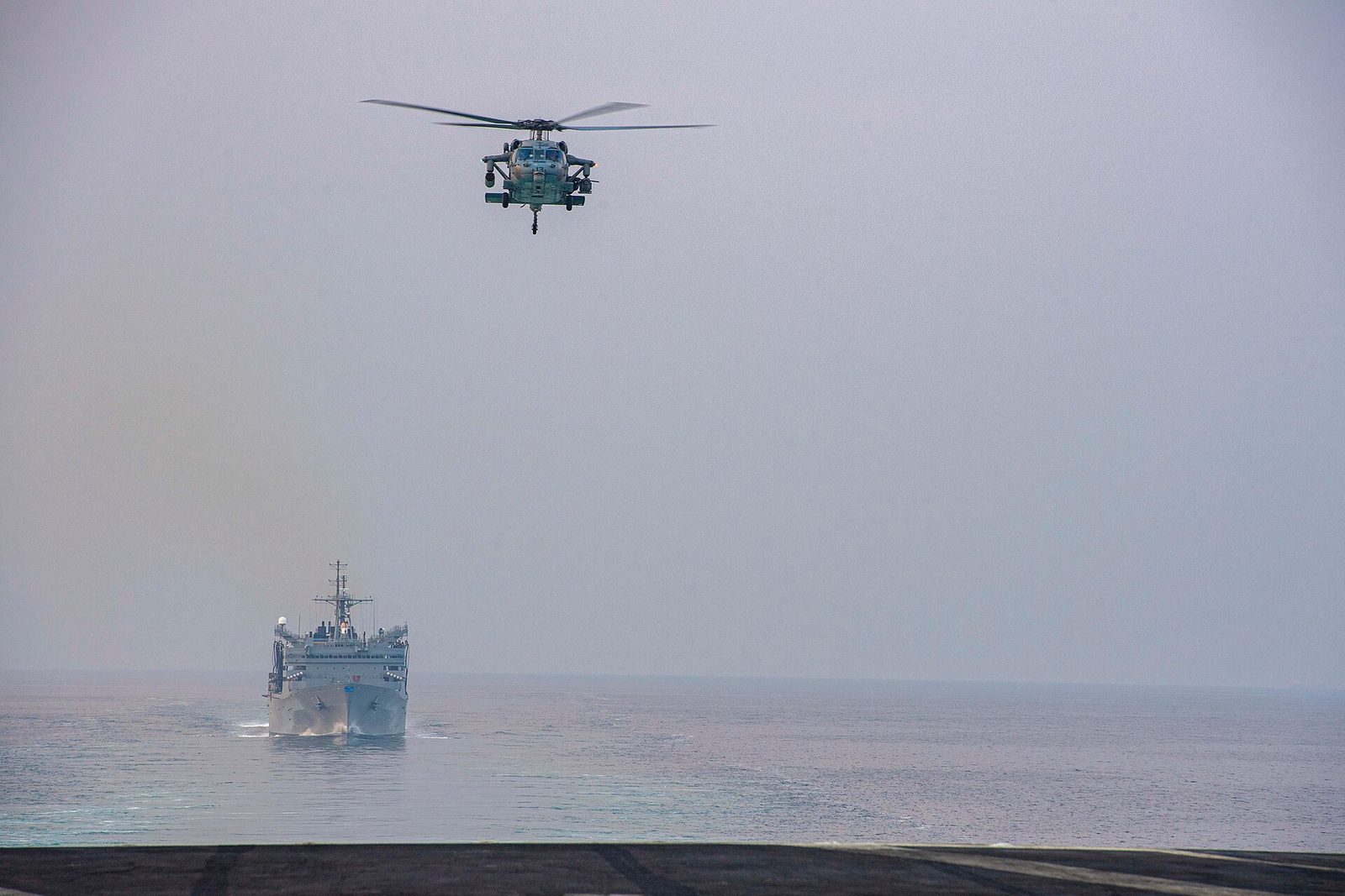 Helicopter hovers over a single ship transiting calm sea, horizon visible, Strait of Hormuz