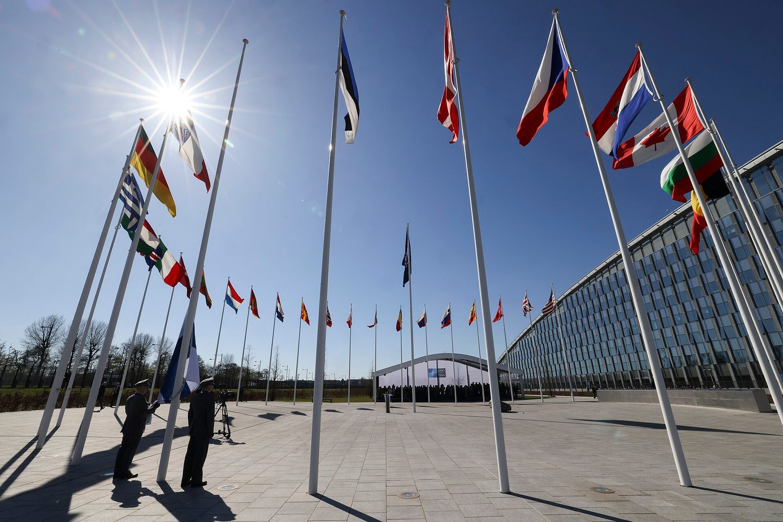 Flagpoles with many national flags outside NATO headquarters, sunlit plaza, Brussels