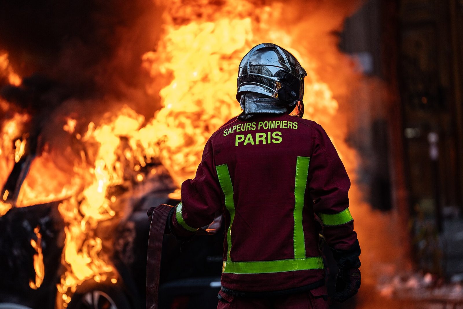 firefighter facing large burning car, 'PARIS' visible on jacket