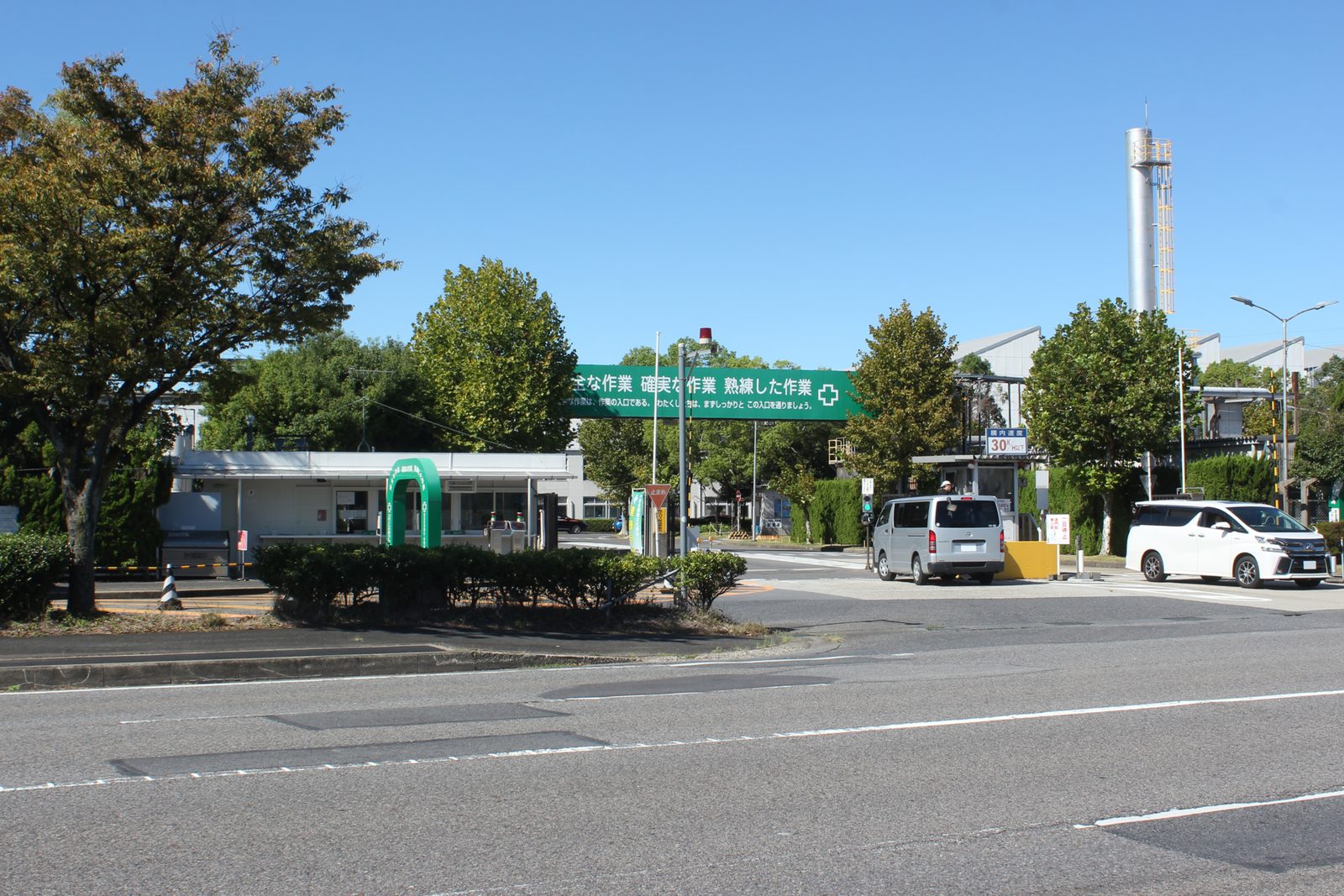factory entrance, trees, parked cars, green safety banner above gate, Takaoka