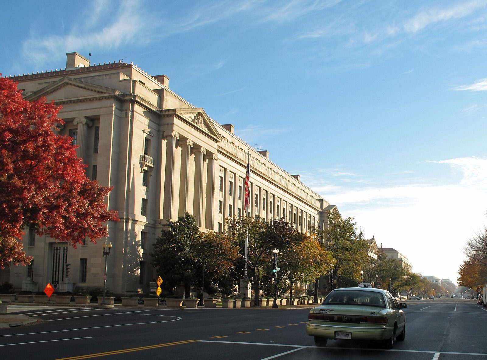 Large neoclassical government building, red-leaved tree, car on quiet road