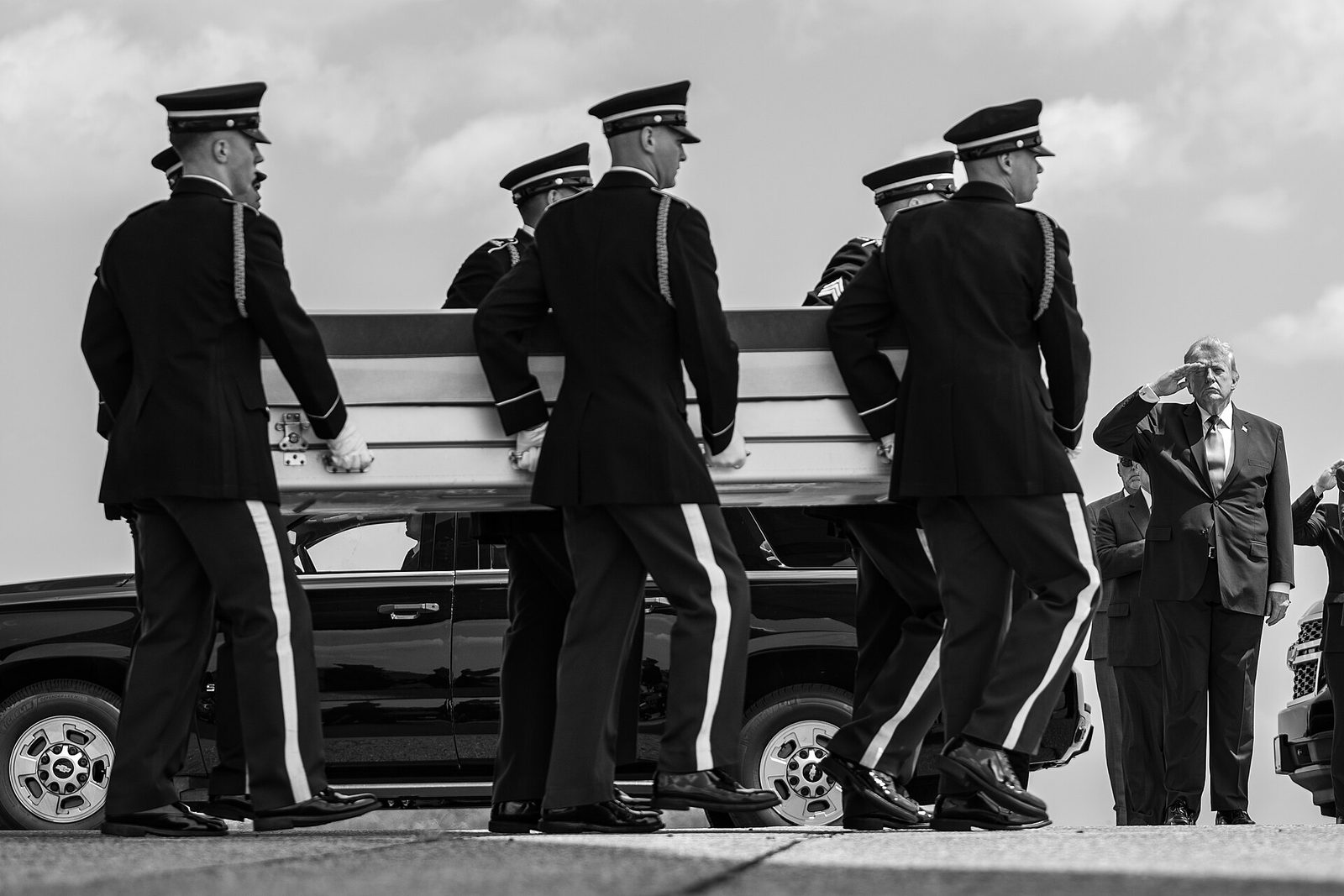 pallbearers carry a coffin, a suited man at right salutes