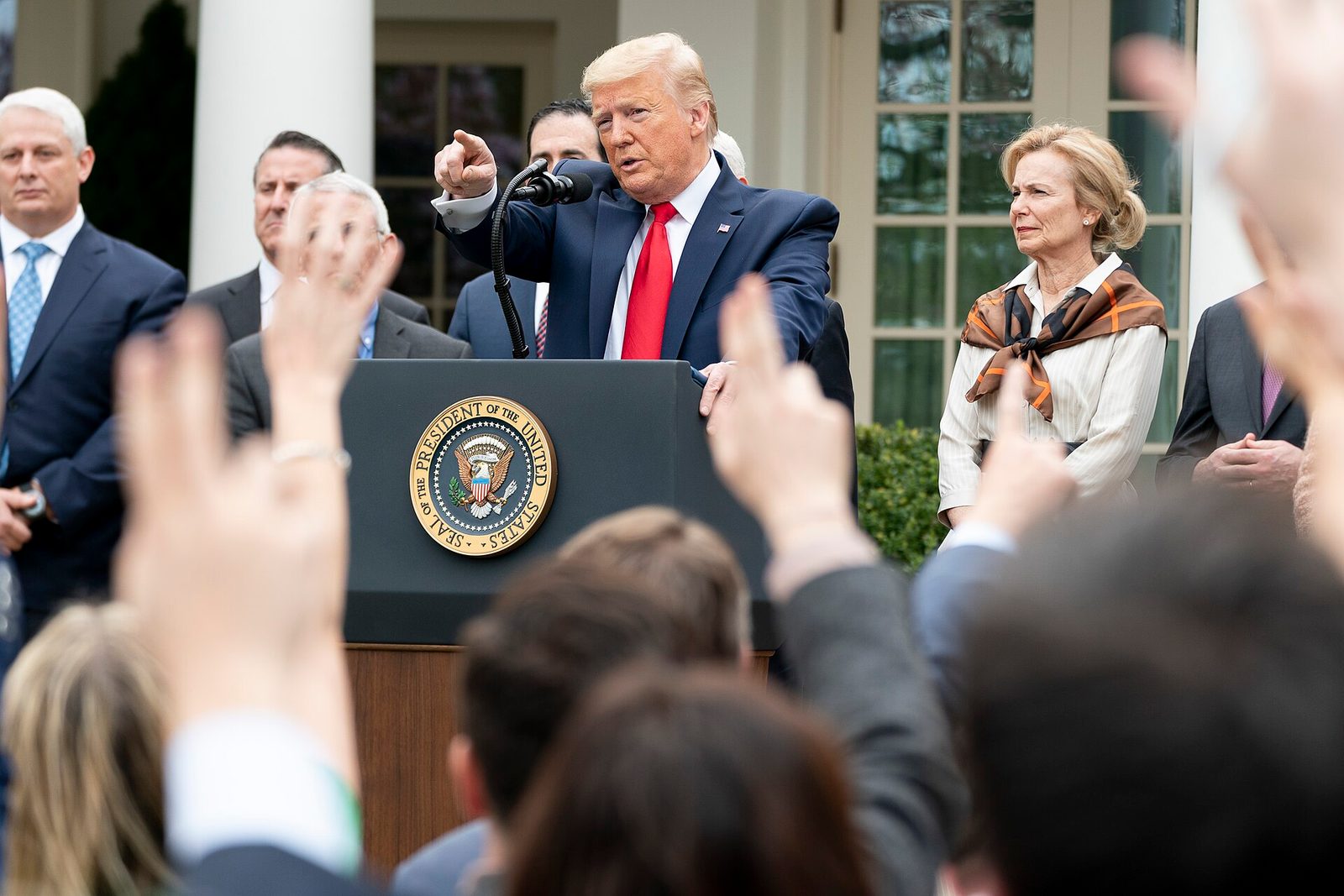 Man at a lectern outdoors, pointing toward raised hands from the crowd