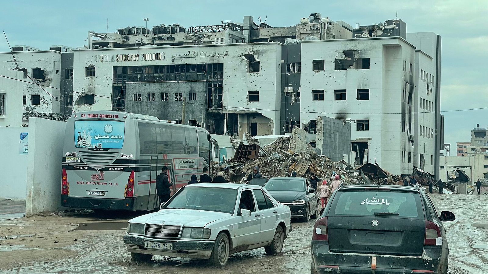 Heavily damaged hospital façade, rubble piled, buses and cars in front, Al-Shifa Hospital