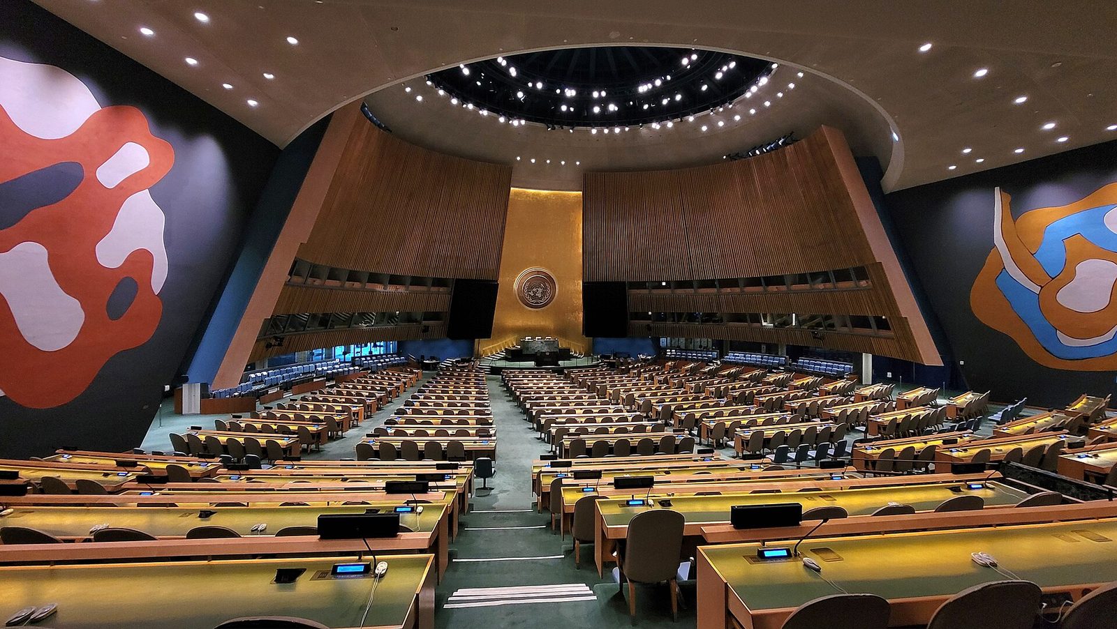 Large semicircular assembly hall with many desks, central podium and mural, United Nations General Assembly Hall