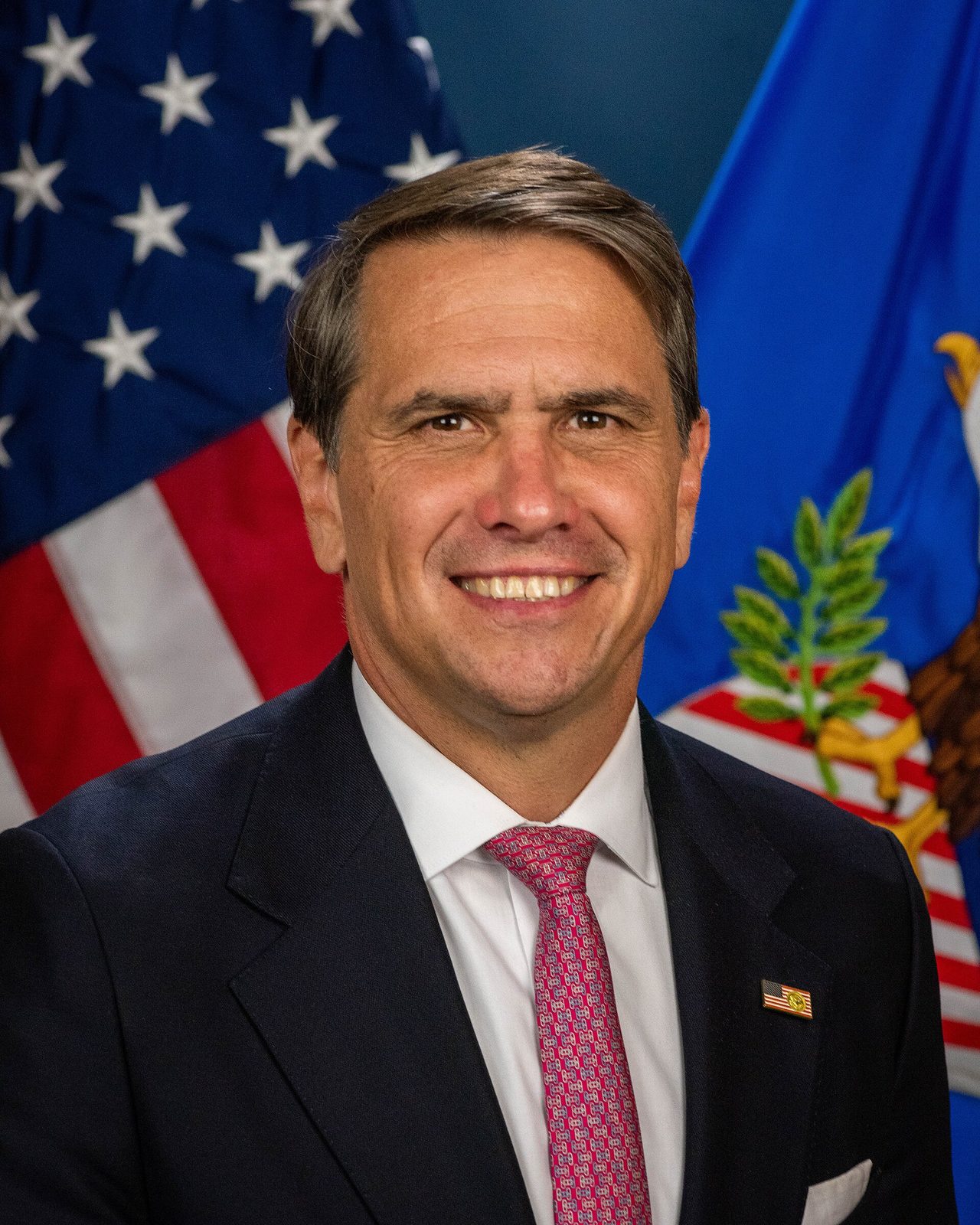 Head-and-shoulders portrait of a man with American flag and seal behind him