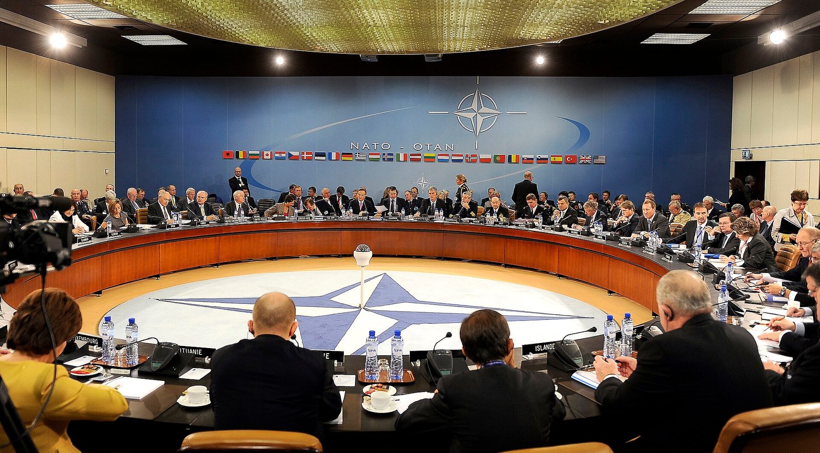 Large meeting room with round table, delegates, NATO emblem on floor, Brussels