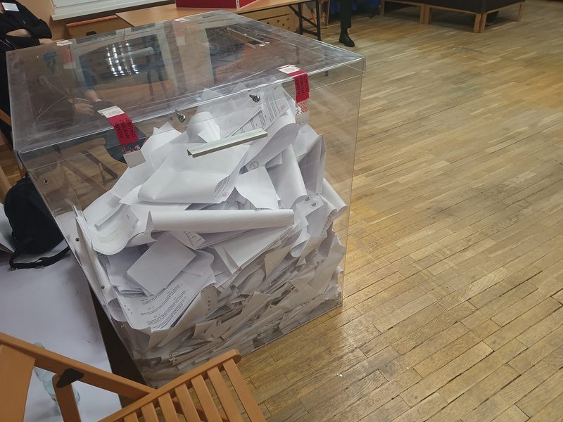 Transparent ballot box filled with folded ballots on wooden floor