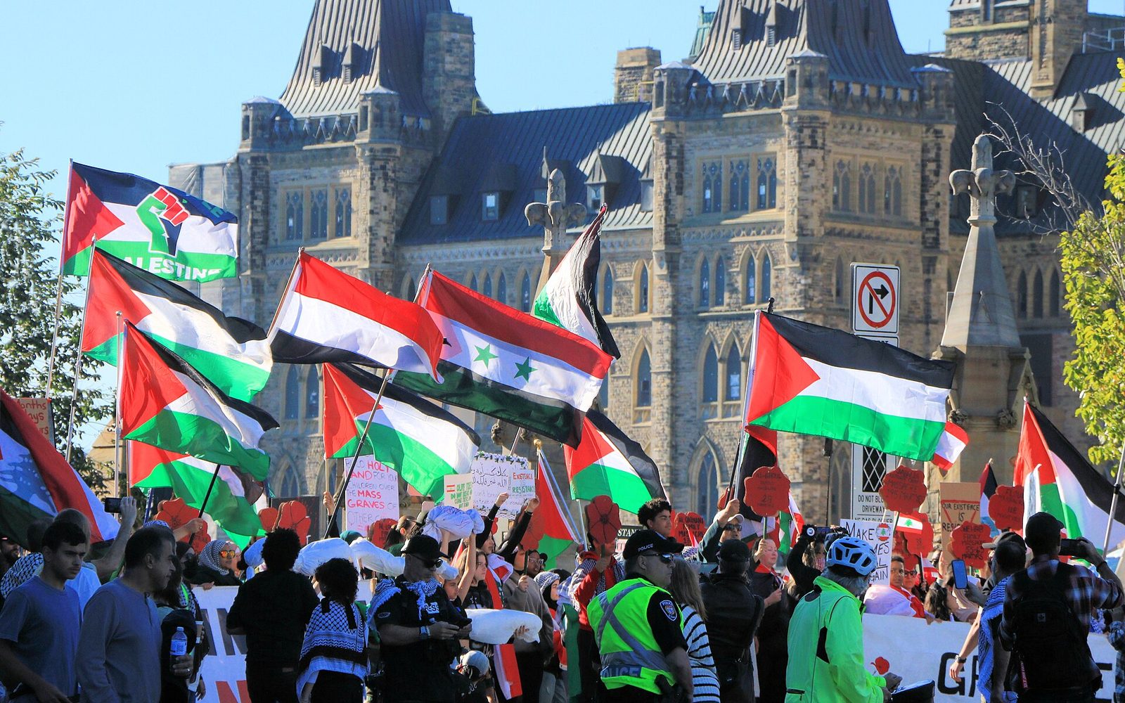 Crowd waving Palestinian flags and signs, stone government building behind, Canada