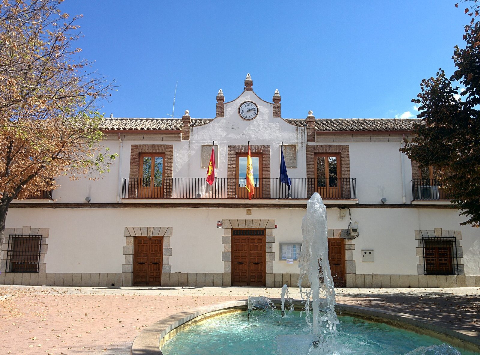 white town hall building, three flags on balcony, fountain foreground, Urda