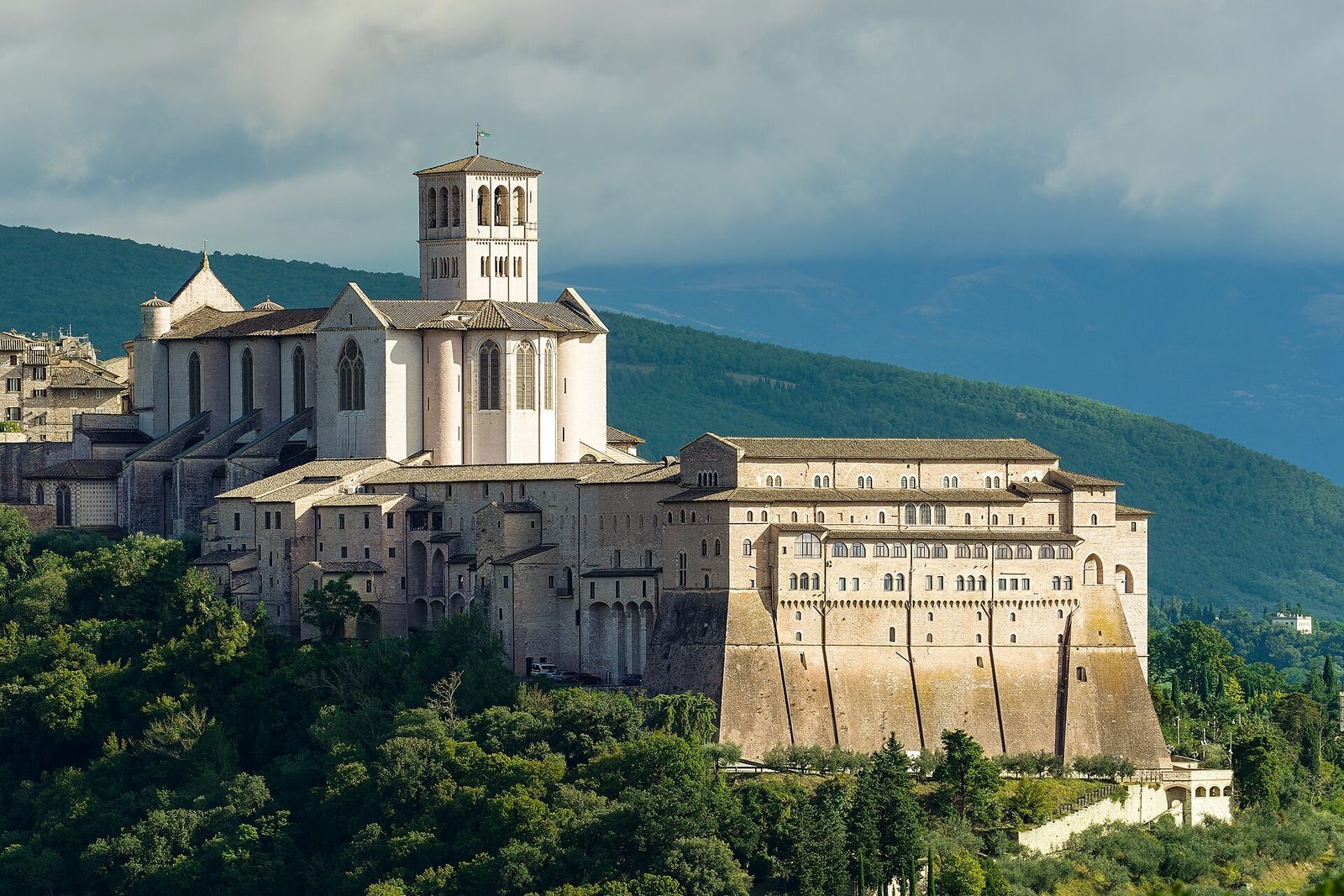 Exterior view of a large basilica on a hillside, trees below, Assisi