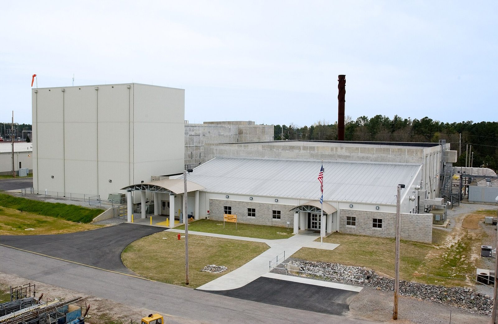 Light-coloured industrial buildings, US flag on pole, paved driveway, surrounding trees