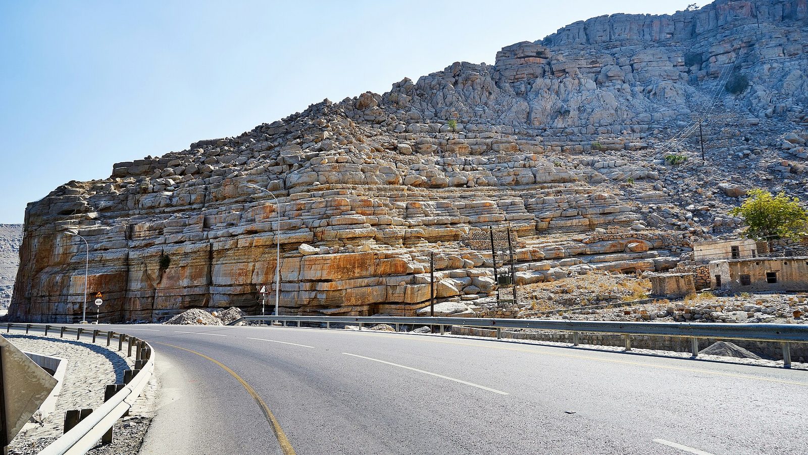 Coastal road curving past layered rocky cliffs, clear sky, Khasab