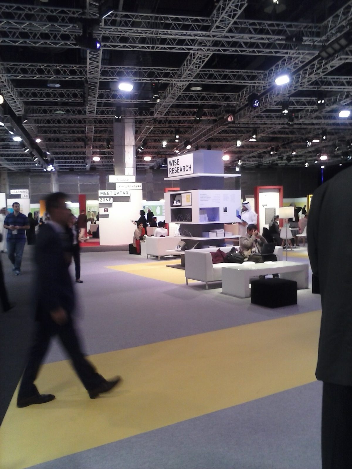 Large exhibition space, 'WISE RESEARCH' sign, people walking and seated, Doha