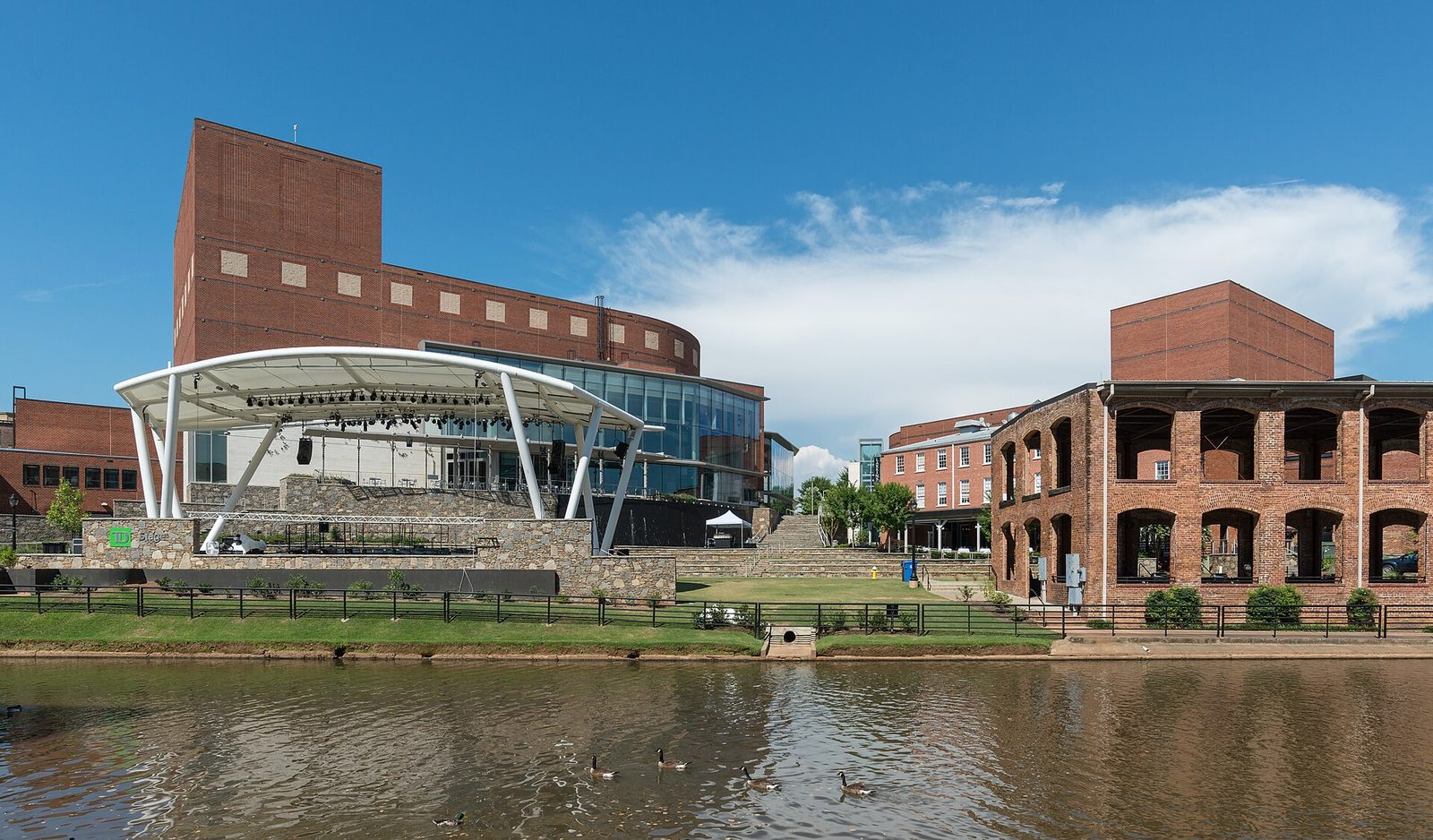 Riverfront performance venue with white stage canopy and brick buildings, Greenville