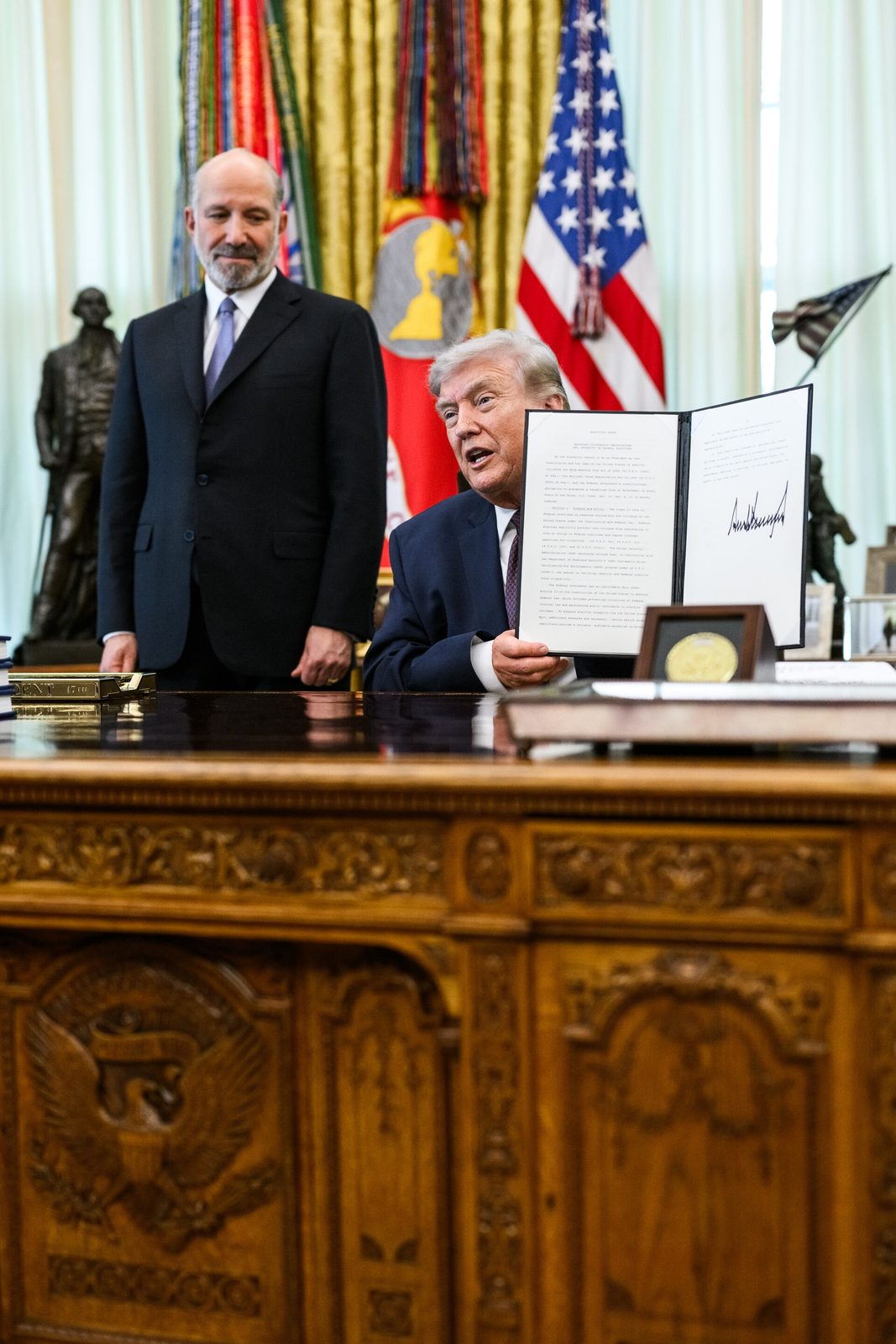 Person at Oval Office desk holds up signed document, standing official behind
