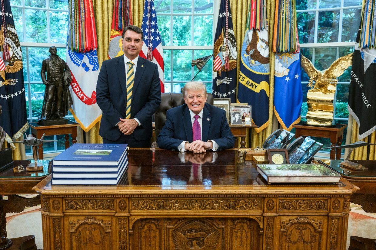 Two men in suits, one seated behind an ornate desk, flags